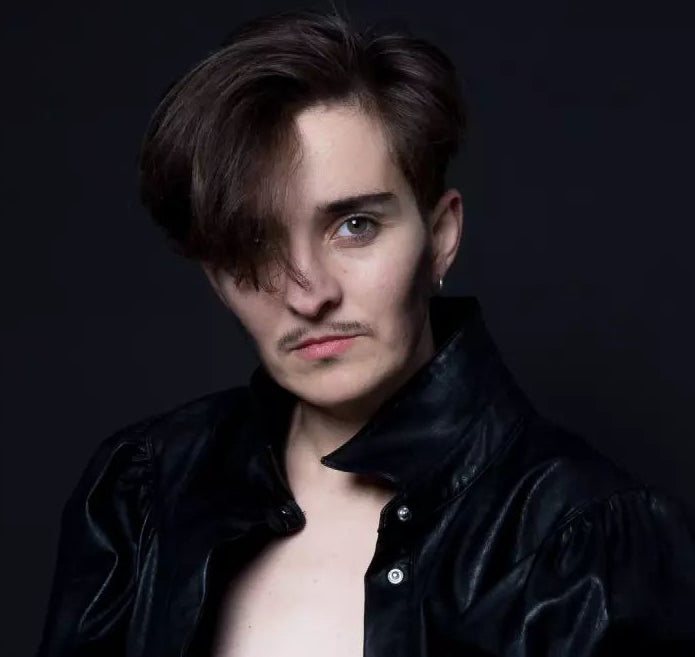 A person with a masculine contour and a drawn-on mustache. They are wearing a black shirt on a black background.